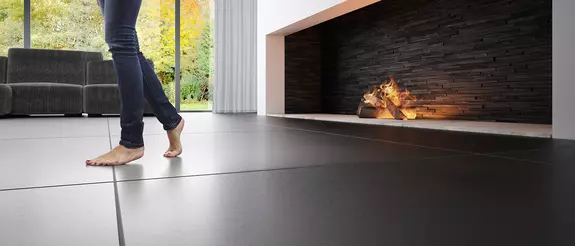 Te zien zijn de benen van een vrouw die in een moderne woonkamer voor een brede kachel op blote voeten over een vloer met grote, donkere tegels loopt.