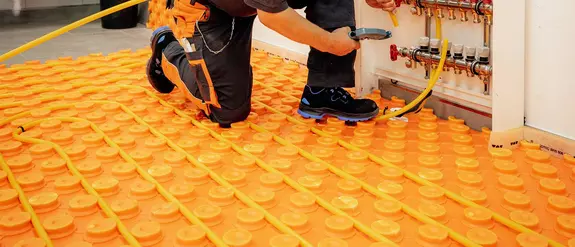 Un profesional corta un tubo de calefacci&oacute;n a la longitud requerida para el circuito de calefacci&oacute;n y lo coloca en una placa de n&oacute;dulos BEKOTEC de Schl&uuml;ter-Systems de color naranja.