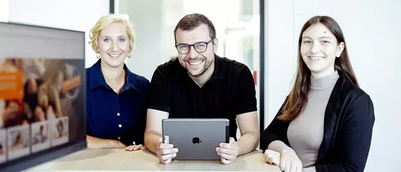Trois employ&eacute;s de Schl&uuml;ter-Systems sont debout autour d’une table, celui du milieu tient une tablette dans ses mains.