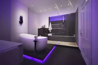 Modernes Bad mit freistehender Badewanne auf einem lila beleuchteten Sockel und offener Dusche