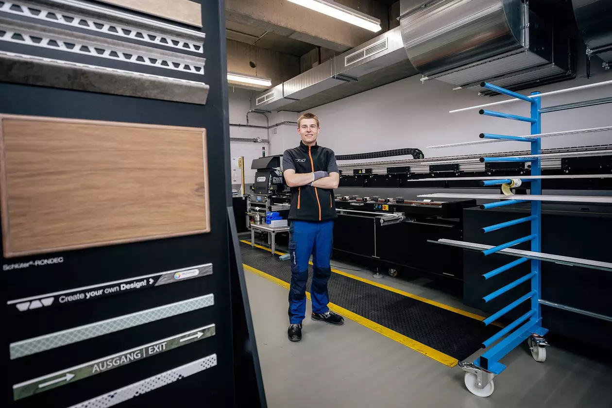 Azubi steht l&auml;chelnd vor Maschine, links ein Aufsteller mit beschichteten Schl&uuml;ter-Systems Profilen