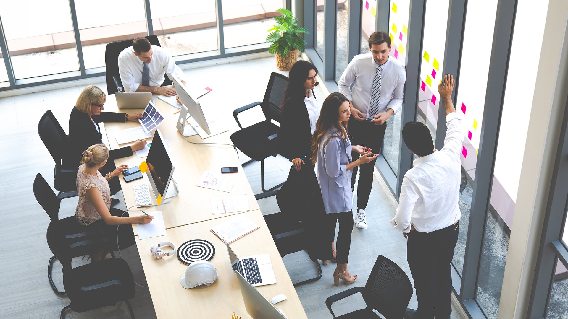 Professionals collaborate around a table and stand pointing to sticky notes on the windows during a team brainstorming session