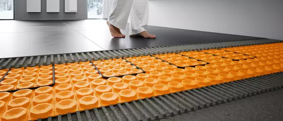 Vrouw loopt over tegelvloer. Daarvoor als doorsnedetekening de eronder gelegde vloerverwarming Schl&uuml;ter-DITRA-HEAT-E.