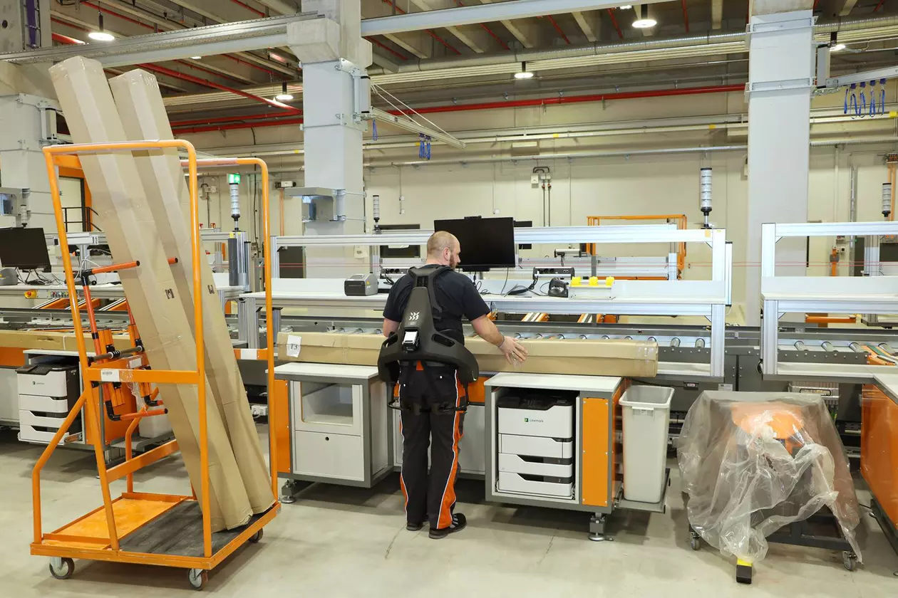 Un collaborateur masculin de Schl&uuml;ter-Systems soul&egrave;ve des marchandises &agrave; l’aide d’un exosquelette devant une ligne d’emballage.