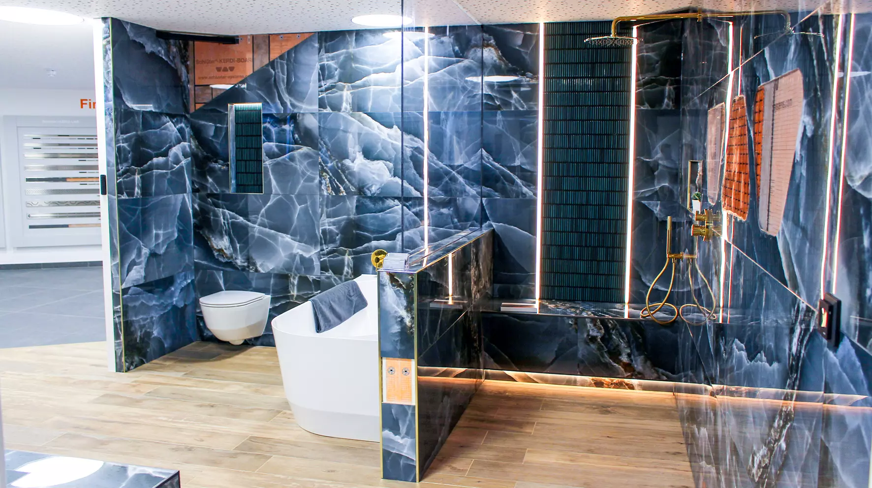 A bathroom has been installed in a showroom setting. On the right, a wall hung toilet and a bath are featured, to the left is a walk-in shower. The tile is blue marble and features cutaways to show the Schl&uuml;ter product