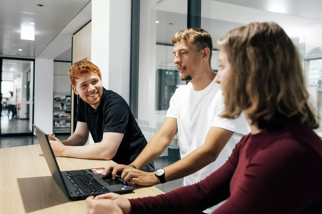 Drei Azubis stehen l&auml;chelnd, an einem Laptop arbeitend, an einem Tisch