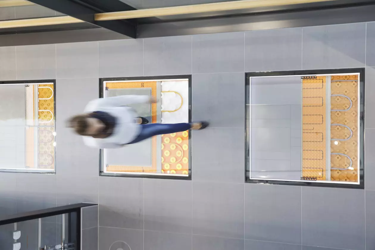 Vue d'en haut sur une femme marchant sur un sol carrel&eacute; o&ugrave; des surfaces vitr&eacute;es laissent entrevoir diff&eacute;rents syst&egrave;mes de chauffage Schl&uuml;ter-Systems dans le support