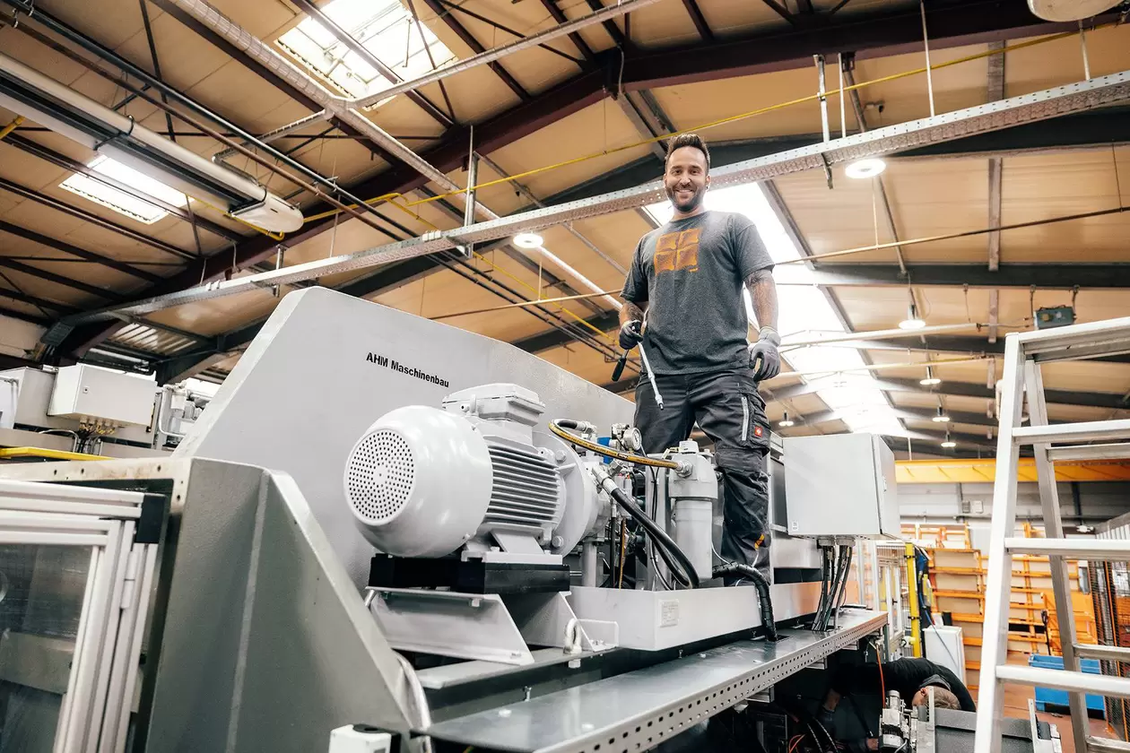 Mitarbeiter steht mit Werkzeug in der Hand auf einer Maschine, um diese zu reparieren und schaut l&auml;chelnd in die Kamera