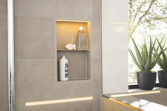 Beige-grey tiled shower wall with a warmly lit, vertical niche for shampoo bottles and toiletries