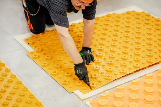 Zu sehen sind drei verschiedene orangefarbene BEKOTEC Estrichnoppenplatten auf einem grauen Fliesenboden liegend, wovon eine gerade mit einem Cuttermesser zurechtgeschnitten wird.
