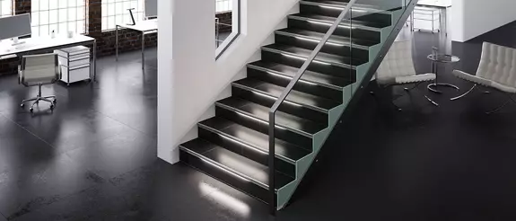 Gro&szlig;z&uuml;giges B&uuml;roloft mit wei&szlig;en Lounge-M&ouml;beln, das in der Mitte durch eine wei&szlig;e Trennwand geteilt ist, vor der eine beleuchtete Treppe mit Metallgel&auml;nder nach oben f&uuml;hrt.
