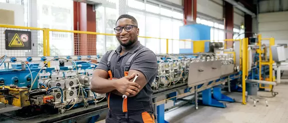 Azubi steht, einen Drehschl&uuml;ssel in der Hand, vor einer Produktionsmaschine und l&auml;chelt in die Kamera