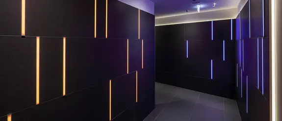 Blick in einen geschwungenen Gang mit schwarz gefliesten W&auml;nden und bunt leuchtenden Lichtlinien in den Fugen