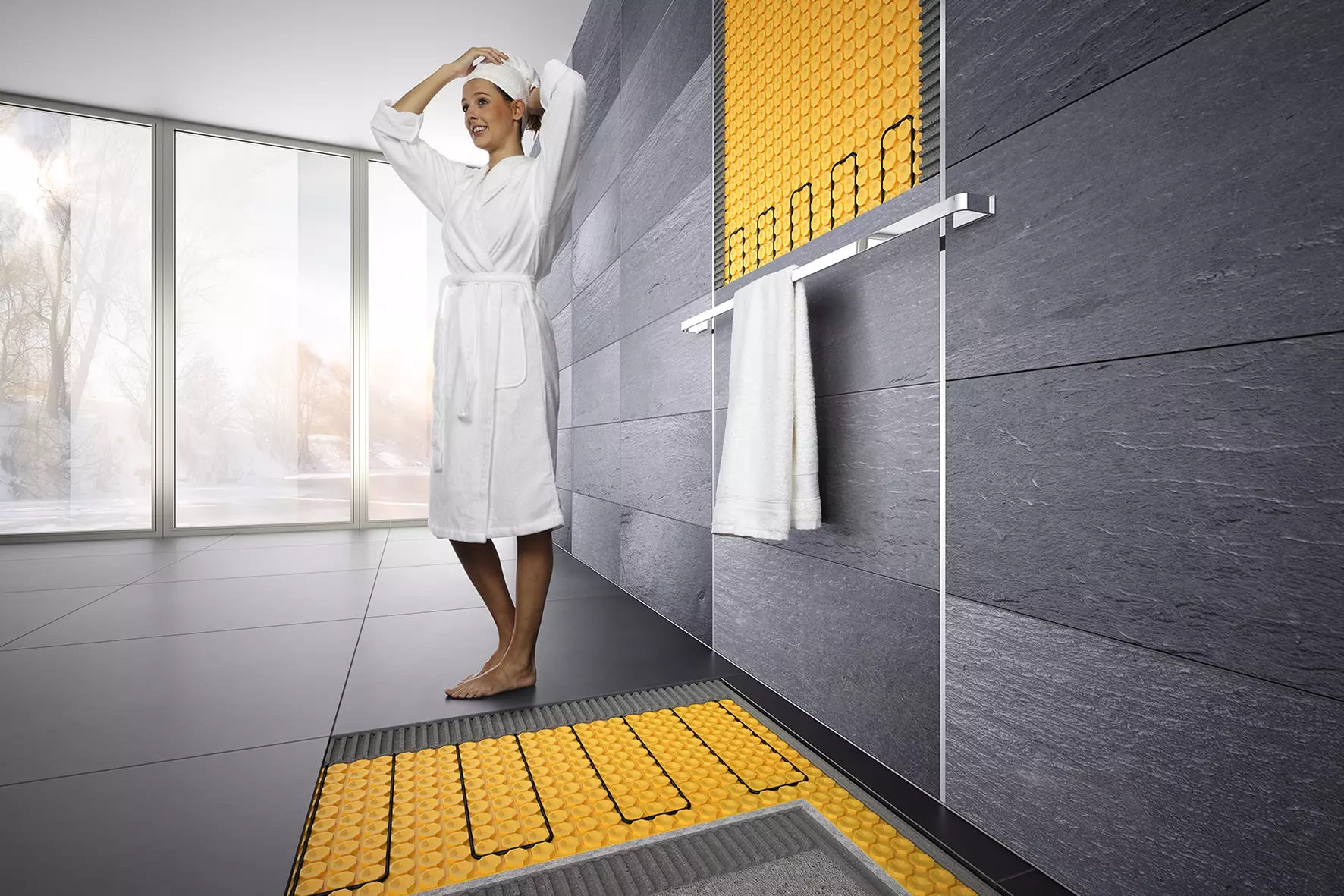 Una joven en albornoz en el cuarto de ba&ntilde;o; la pared de la ducha y el suelo est&aacute;n calefactados con DITRA-HEAT-E.