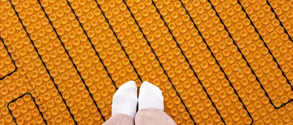 Two women's feet in white socks stand on an orange Schl&uuml;ter-DITRA-HEAT mat with black heating cables