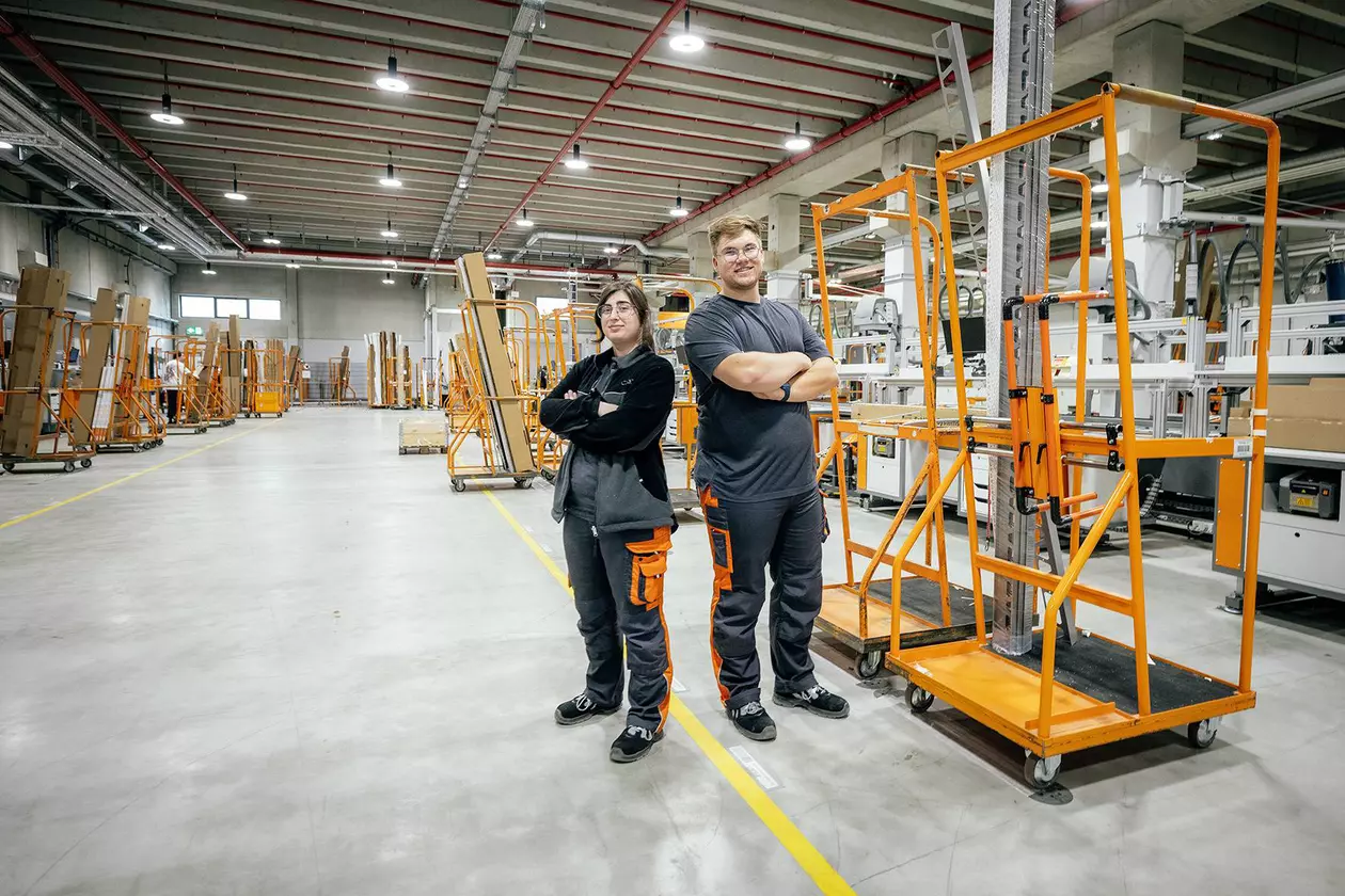 Zwei Auszubildende stehen in ausgeleuchteter Logistik-Halle, im Hintergrund Pakete
