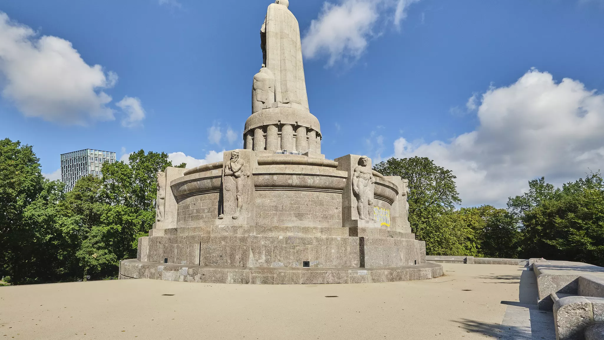 Bismarck-monumentet p&aring; en stor plats, figurens rygg syns framf&ouml;r en bl&aring; himmel med n&aring;gra moln och gr&ouml;na tr&auml;dkronor.