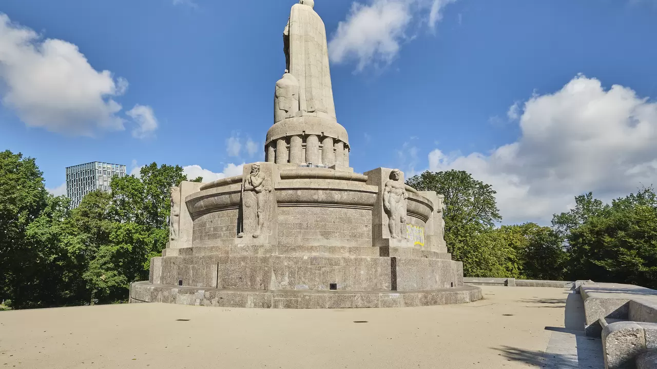 Bismarck-monumentet p&aring; en stor plats, figurens rygg syns framf&ouml;r en bl&aring; himmel med n&aring;gra moln och gr&ouml;na tr&auml;dkronor.