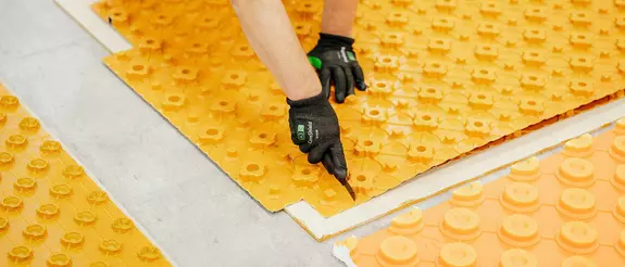 En la imagen se pueden ver tres placas de n&oacute;dulos BEKOTEC diferentes de color naranja sobre un suelo de baldosas grises, una de las cuales se est&aacute; cortando con un c&uacute;ter.