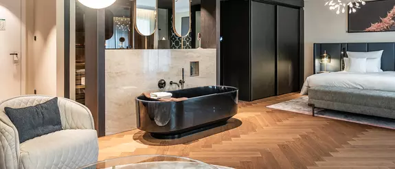 Moderna habitaci&oacute;n del hotel Steigenberger Hof, con iluminaci&oacute;n c&aacute;lida, suelo laminado, cama tapizada y ba&ntilde;era exenta de color negro