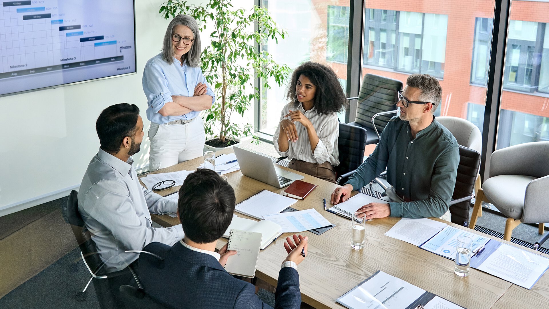 A corporate development team collaborates in a meeting room with a Gantt chart visible on a screen in the background.