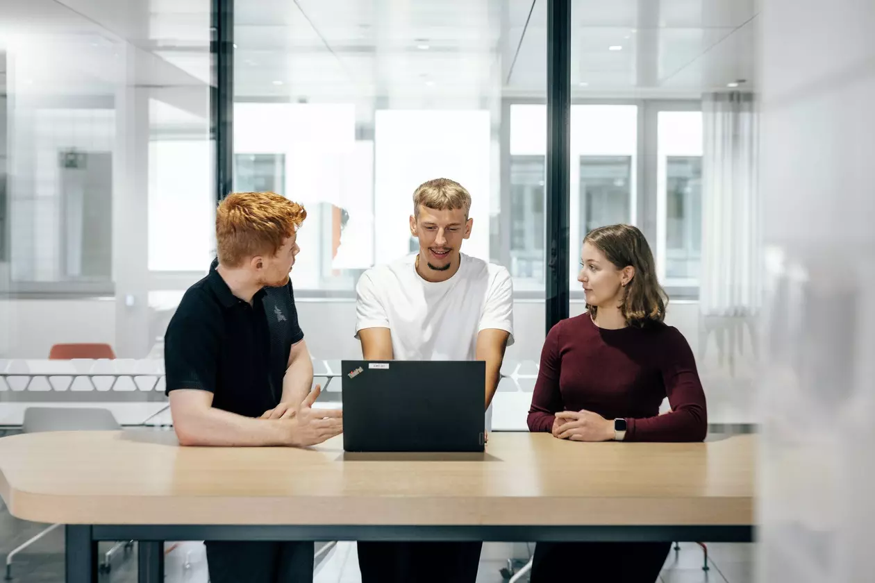 Drei Azubis stehen an einem Tisch, blicken auf einen Laptop und unterhalten sich