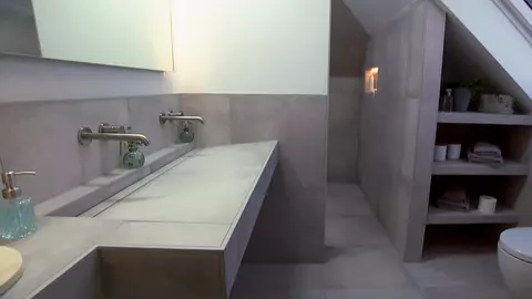 Blik in een badkamer met schuin plafond, dubbele wastafel, betegeld inbouwrek en verlichte wandnis