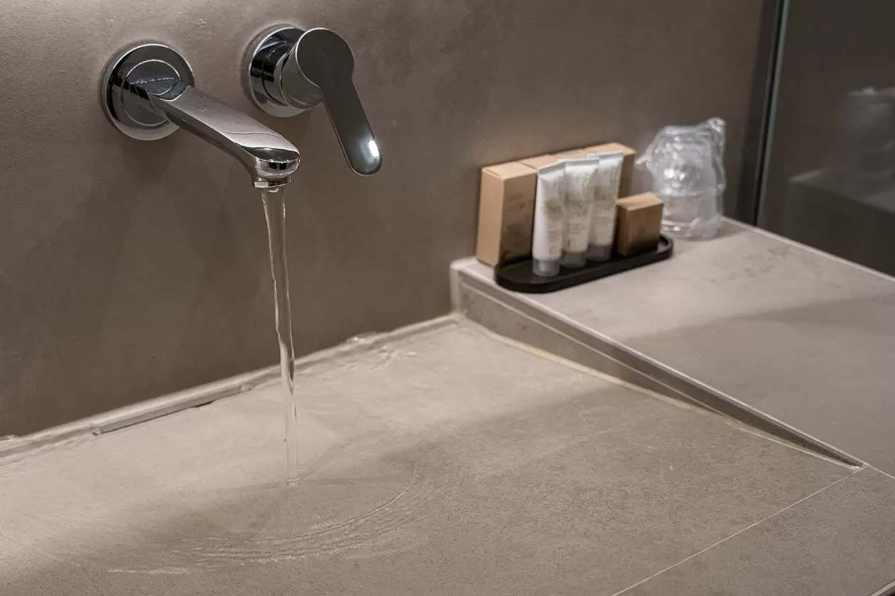 Silberne Armatur mit flie&szlig;endem Wasser &uuml;ber einem Waschtisch. Die verbauten Profile Schl&uuml;ter-JOLLY und KERDI-LINE-VARIO in steingrauer Strukturbeschichtung f&uuml;gen sich harmonisch in die beige Fliesenoptik ein. Auf dem Waschtisch sind einige Waschartikel. 