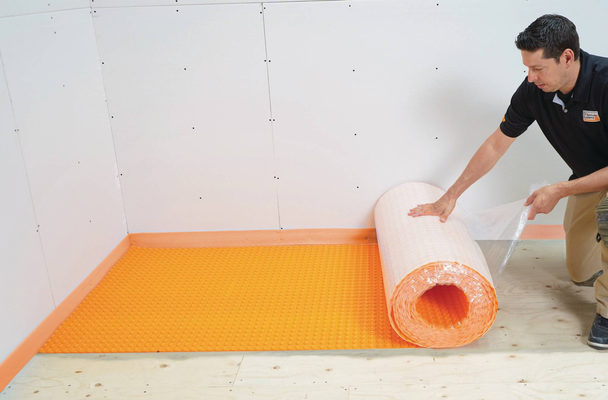 Un homme installe un rouleau de membrane orange autocollante Schluter sur un plancher de contreplaqué