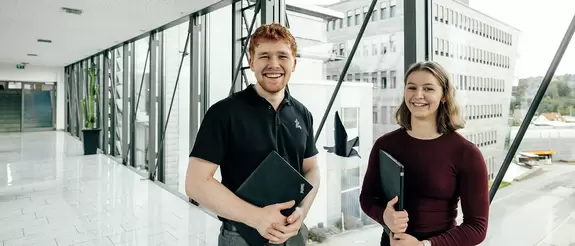 Azubi und Azubine schauen l&auml;chelnd, ihre Arbeitslaptops unter dem Arm, in die Kamera