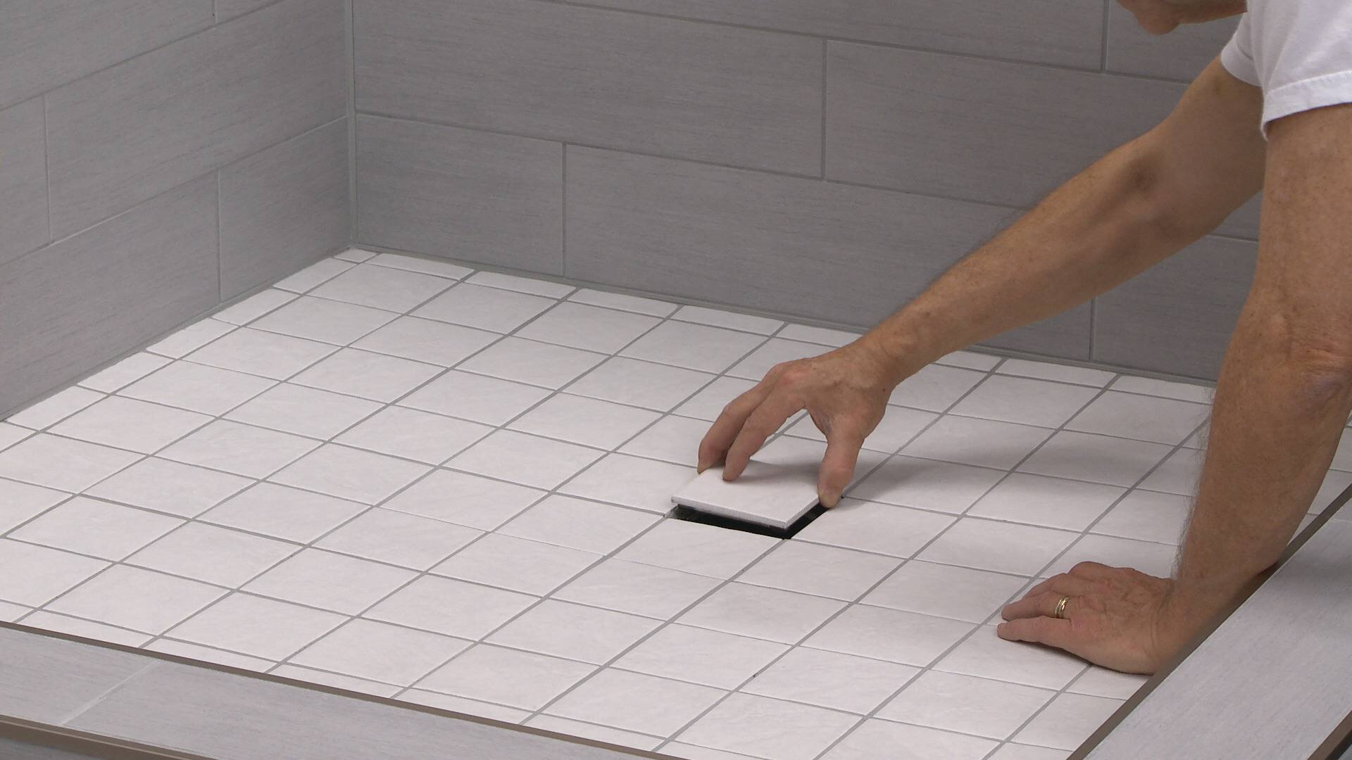 Une personne pose le dernier carreau carré sur un sol de douche carrelé proprement fini, mettant en valeur une grille de drain carrelée.