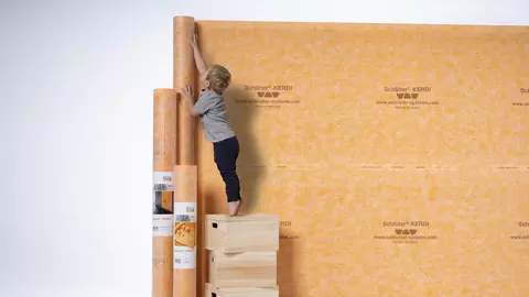 Ein kleiner Junge rollt orange-farbige KERDI-Abdichtungsbahnen an einer Wand ab.
