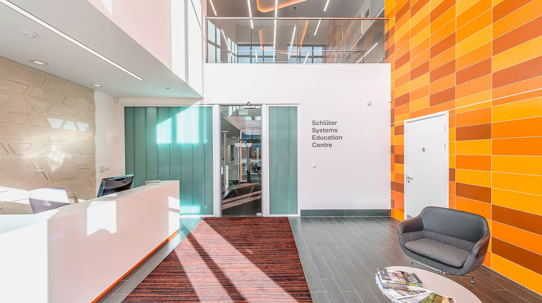 The entrance foyer to Schl&uuml;ter-Systems Ltd featuring a doorway into the building, a reception desk to the left and seating area on the right. Behind the seating area, a wall with orange tile, heated with DITRA-HEAT
