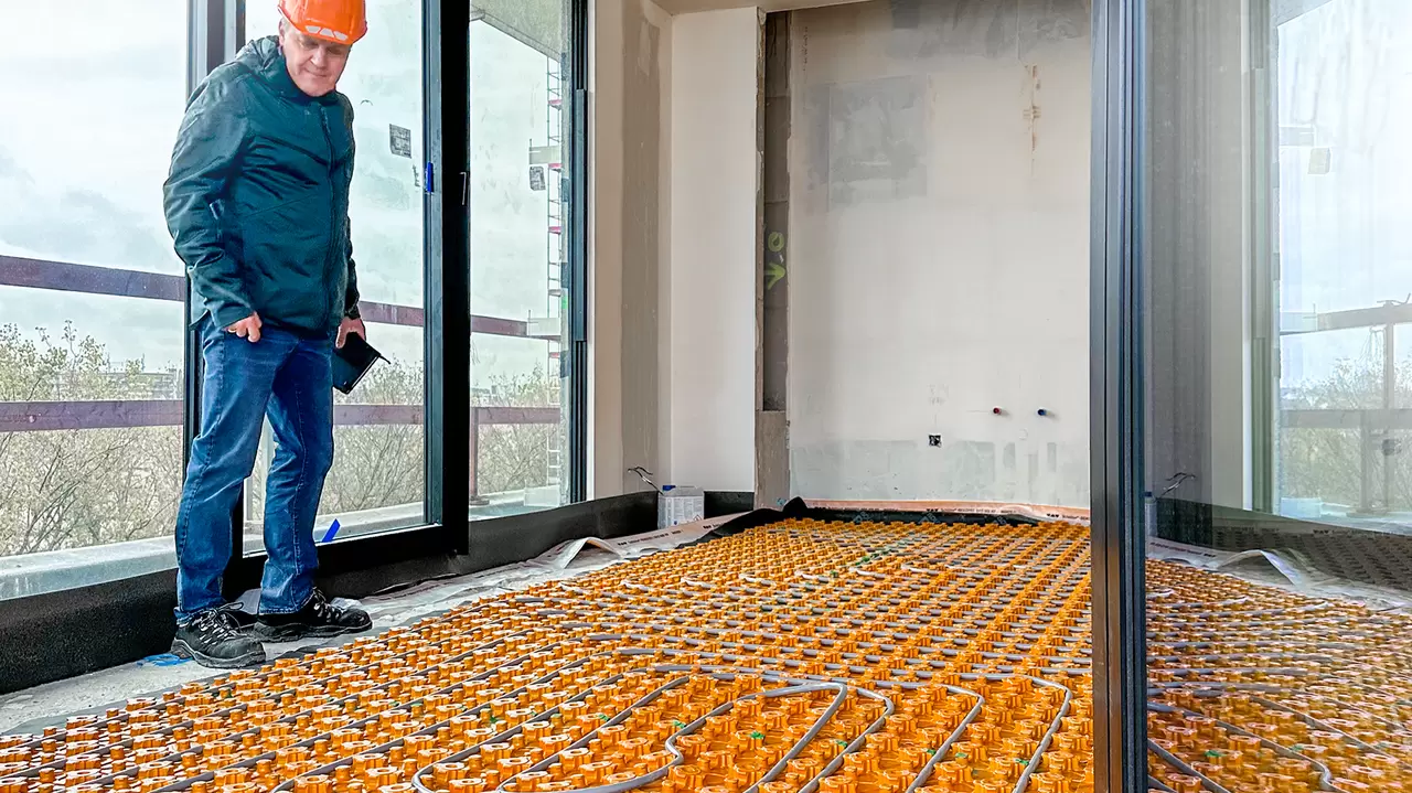 Een man met een veiligheidshelm bekijkt kritisch de gelegde BEKOTEC-THERM-mat met verwarmingsbuizen in een ruimte van City Dox
