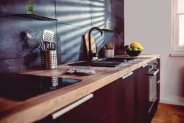 Cuisine moderne avec carrelage fonc&eacute;, plan de travail en bois, appareils &eacute;lectrom&eacute;nagers noirs, robinetterie noire et deux tablettes design Schl&uuml;ter-SHELF en noir mat au mur.