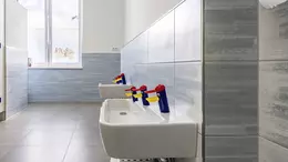 Wei&szlig;e Waschbecken mit  bunten, kindgerechten Wasserh&auml;hnen vor einer hellgaru, gro&szlig;formatig gefliesten Wand in einem Kindergarten