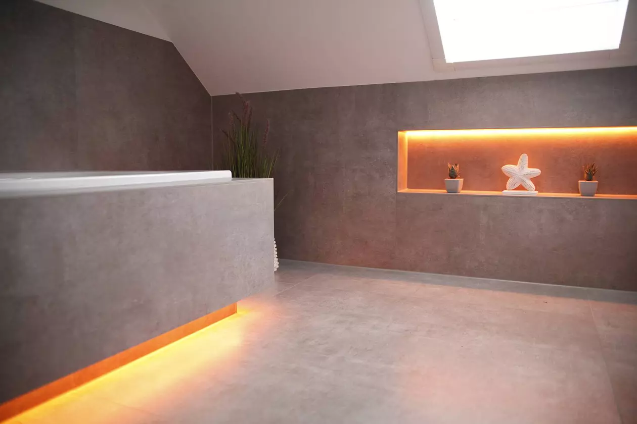 Ein beige-grau gefliestes Badezimmer mit Dachschr&auml;ge und warmer Beleuchtung an Badewanne und Nische