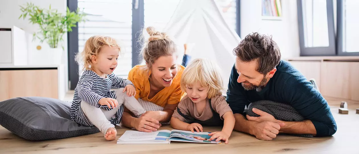 Un jeune couple, une femme et un homme, avec deux jeunes enfants, est allong&eacute; sur le sol et feuillette un livre pour enfants en souriant.