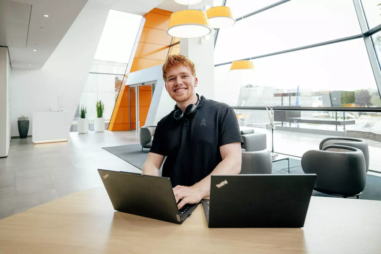 Azubi, mit Headset um den Hals, steht hinter zwei aufgeklappten Laptops und blickt l&auml;chelnd in die Kamera