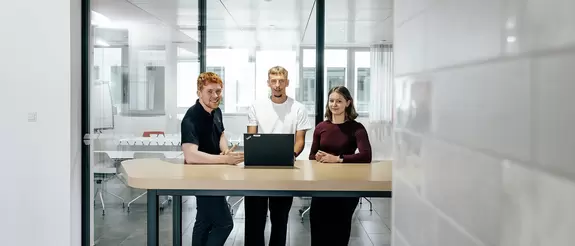 Drei Azubis stehen hinter einem Laptop an einem Tisch und blicken in die Kamera