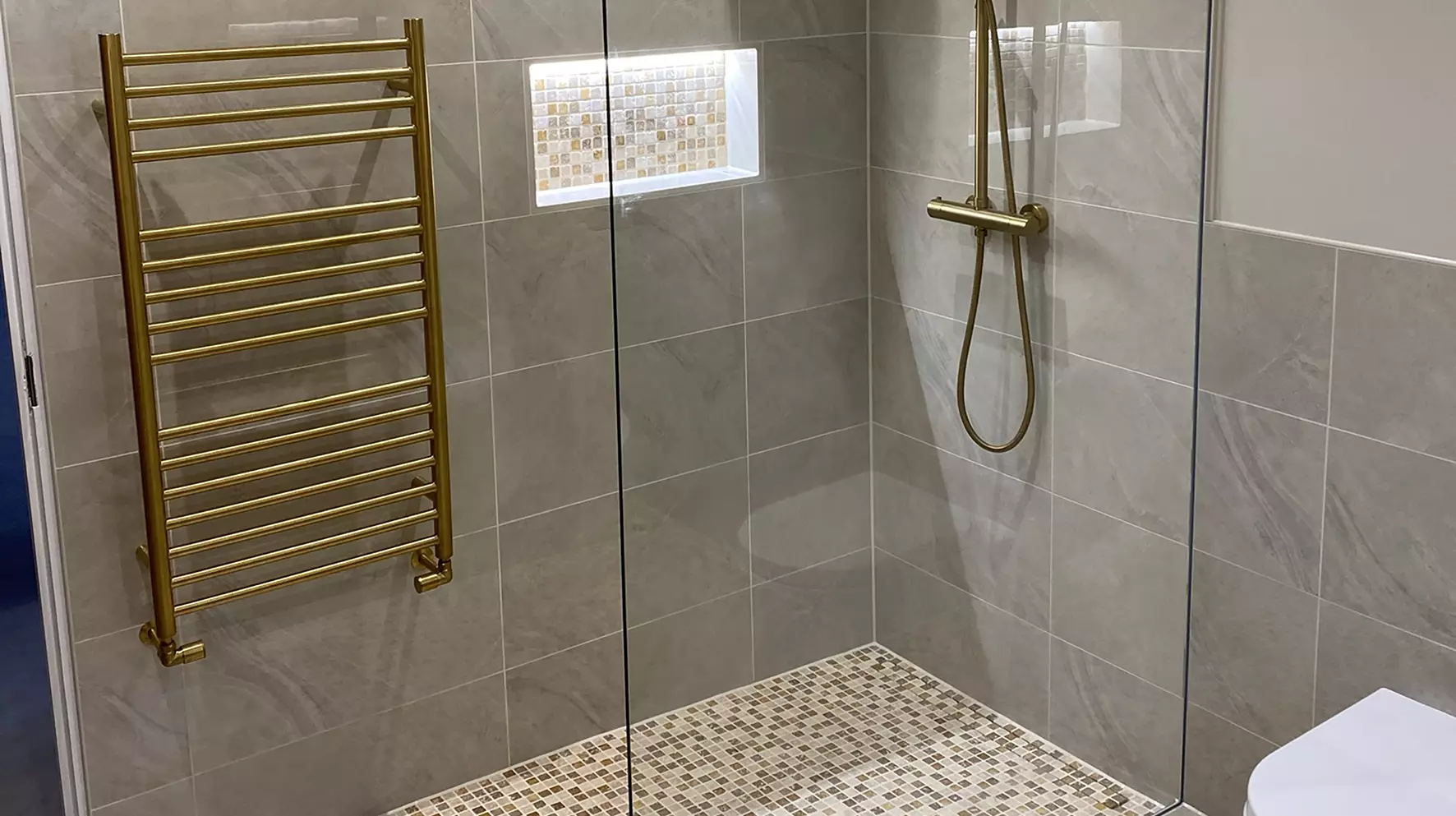 Ducha a nivel de suelo con suelo de mosaico, hornacina a juego en la pared, radiadores dorados y grifer&iacute;a de ducha.