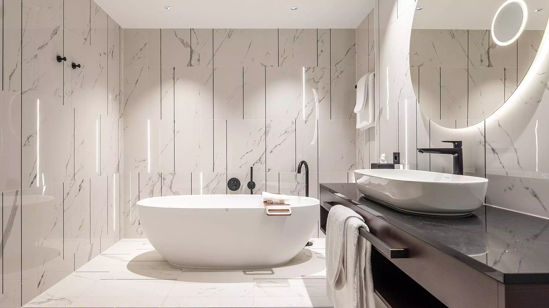 Cuarto de ba&ntilde;o de gran tama&ntilde;o con baldosas de m&aacute;rmol blanco, iluminaci&oacute;n en las paredes y ba&ntilde;era exenta