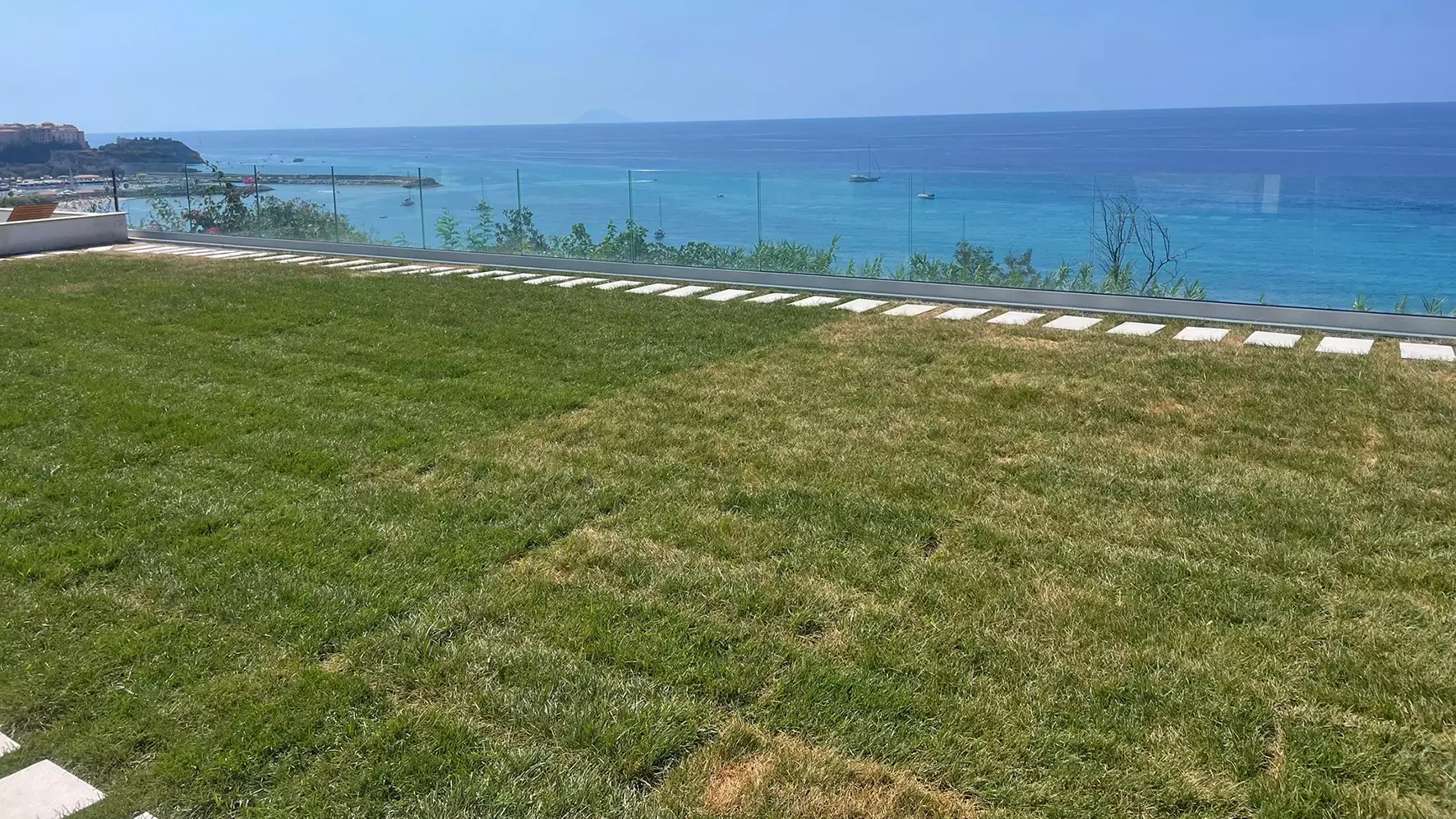 Terrazza sul tetto con prato e balaustra in vetro di fronte a un panorama costiero con vista sul mare